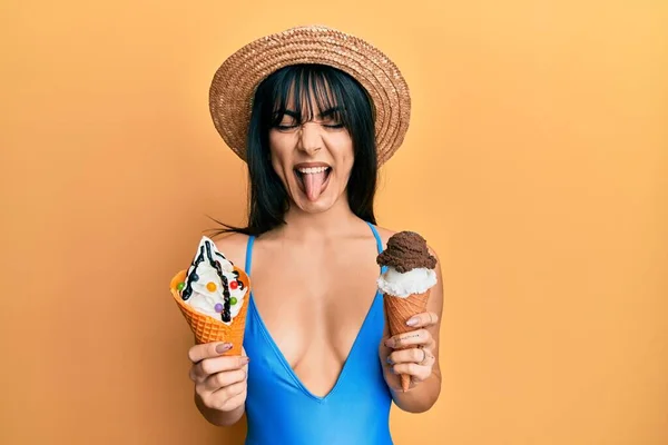 Young brunette woman with bangs wearing swimwear holding two ice cream cones sticking tongue out happy with funny expression.