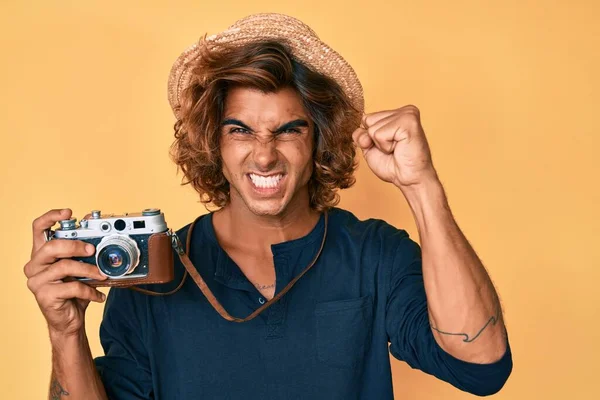 Young hispanic man wearing hat holding vintage camera annoyed and frustrated shouting with anger, yelling crazy with anger and hand raised