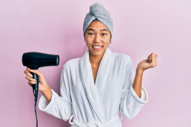 Duş havlusu şapkası ve bornoz giyen genç Çinli kadın kurutucuyu tutarken gururla bağırıyor, zaferi kutluyor ve başarıyı kollarını kaldırarak kutluyor. 