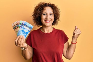 Beautiful middle age mature woman holding canadian dollars screaming proud, celebrating victory and success very excited with raised arm 