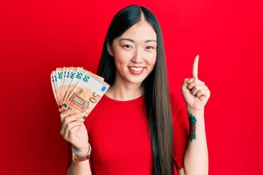 50 Euro 'luk banknotları elinde tutan genç Çinli bir kadın bir fikir veya soruyla gülümsüyor ve mutlu bir yüzle işaret ediyor. 