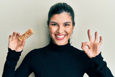 Sağlıklı enerji atıştırmalığı olarak protein yiyen genç İspanyol kadın parmaklarıyla işaret veriyor, gülümseyerek mükemmel bir sembol oluşturuyor. 
