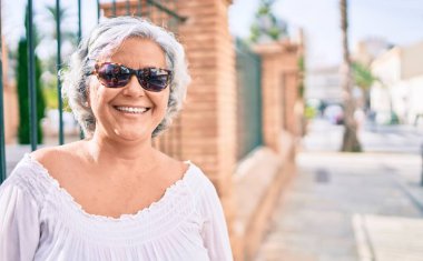 Beyaz saçlı orta yaşlı bir kadın mutlu bir şekilde dışarıda gülümsüyor.