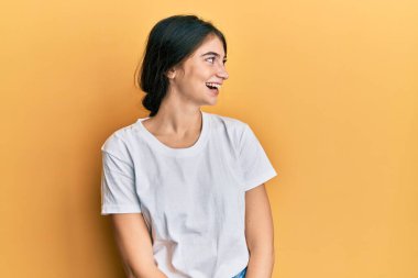 Beyaz gündelik tişört giyen genç beyaz kadın yüzünde gülümseme, doğal bir ifade ile yan yana bakıyor. Kendinden emin gülen. 