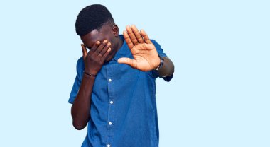 Genç, Afro-Amerikan bir adam, günlük kıyafetler giyiyor, elleriyle gözleri örtüyor ve üzgün ve korku ifadeleriyle jestleri durduruyor. utangaç ve olumsuz kavram. 