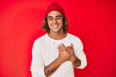 Young hispanic man wearing wool sweater and winter hat smiling with hands on chest with closed eyes and grateful gesture on face. health concept. 