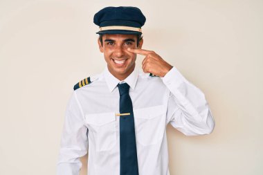 Young hispanic man wearing airplane pilot uniform pointing with hand finger to face and nose, smiling cheerful. beauty concept 