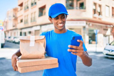 Kurye üniforması giyen Afrikalı kurye dışarıda akıllı telefon kullanıyor.