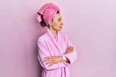 Sarışın, duş başlığı ve bornoz takan genç bir kadın kolları çapraz ve kendinden emin bir şekilde yana bakıyor. 