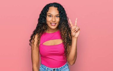 Kıvırcık saçlı, pembe tişörtlü, gülümseyen ve kameraya göz kırpan genç İspanyol kadın zafer işareti yapıyor. İki numara.. 