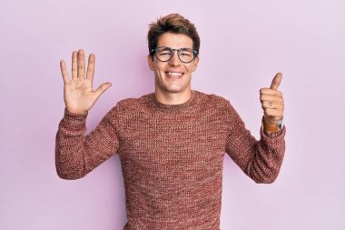 Yakışıklı beyaz adam, sıradan süveter giyiyor ve gözlük takıyor. Altıncı parmaklarını gösterip işaret ediyor. Kendinden emin ve mutlu bir şekilde gülümsüyor.. 