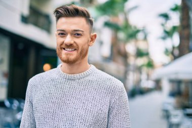 Genç beyaz adam gülümseyerek şehirde mutlu mesut duruyor..