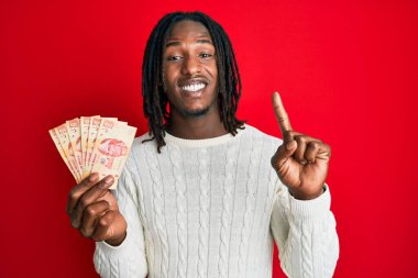 Saçları örgülü Afrikalı Amerikalı bir adam elinde 100 Meksika Pezosu ile bir fikir veya soru işareti ile gülümsüyor. 
