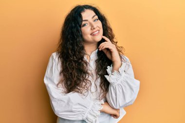 Kıvırcık saçlı, sıradan kıyafetler giyen genç esmer kadın kameraya kolları çapraz ve çenesi açık bakıyor. Olumlu düşünmek. 