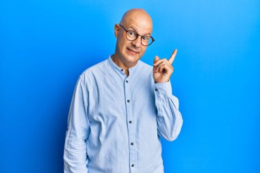 Orta yaşlı kel bir adam, günlük kıyafetler ve gözlük takıyor. Yüzünde kocaman bir gülümseme var. Eli ve parmağı kameraya bakıyor.. 