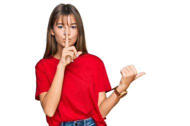 Teenager caucasian girl wearing casual red t shirt asking to be quiet with finger on lips pointing with hand to the side. silence and secret concept. 