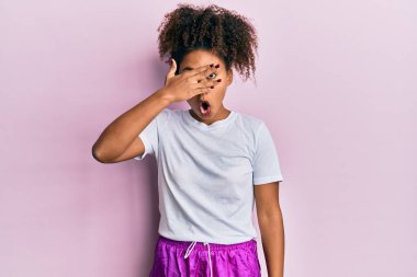 Beautiful african american woman with afro hair wearing sportswear peeking in shock covering face and eyes with hand, looking through fingers with embarrassed expression. 
