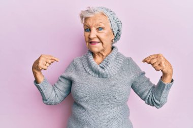 Yaşlı, gri saçlı, yün kazak giyen, kışlık şapkalı, kendine güvenen, gülümseyen, kendini gururlu ve mutlu gösteren bir kadın.. 