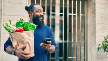 Sakallı Afrikalı Amerikalı adam elinde süpermarketten alınmış kağıt torbalarla akıllı telefon kullanıyor.