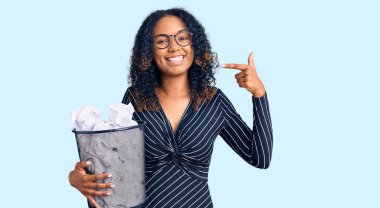 Genç Afrikalı Amerikalı bir kadın elinde buruşuk kağıtlarla dolu bir çöp tenekesiyle kendini mutlu ve gururlu olarak gösteren bir gülümsemeyi işaret ediyor. 