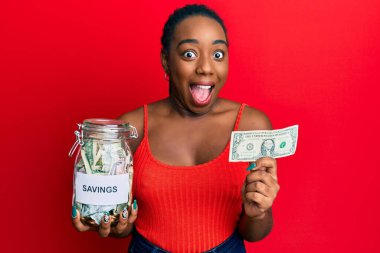 Young african american woman holding jar with savings and 1 dollar banknote celebrating crazy and amazed for success with open eyes screaming excited. 