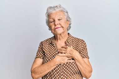 Üstün gri saçlı, günlük kıyafetler giyen, elleri göğsünde, gözleri kapalı gülümseyen ve yüzünde minnet ifadesi olan bir kadın. sağlık kavramı. 