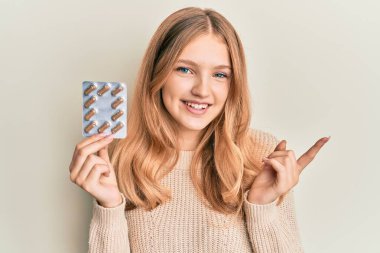 Beautiful young caucasian girl holding pills smiling happy pointing with hand and finger to the side 