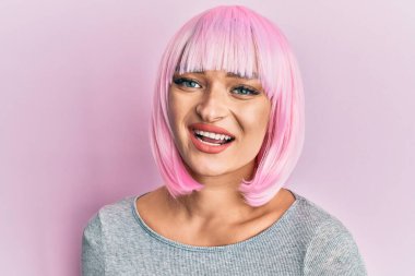 Pembe peruklu genç beyaz kadın pozitif ve mutlu görünüyor. Kendine güvenen bir gülümsemeyle ayakta duruyor ve dişlerini gösteriyor. 