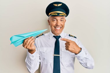 Yakışıklı, orta yaşlı, uçak pilotu üniforması giyen, elinde kağıt uçakla gülümseyen, mutlu, eli ve parmağı olan bir adam. 