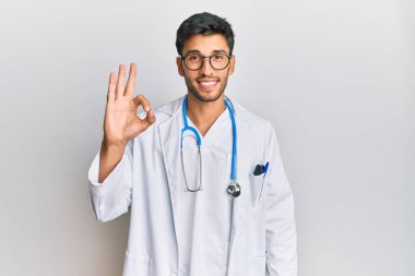 Doktor üniforması ve steteskop giymiş genç yakışıklı adam olumlu gülümsüyor. El ve parmaklarıyla 