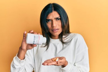 Young latin transsexual transgender woman holding pills clueless and confused expression. doubt concept. 