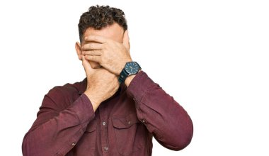 Gündelik kıyafetler giyen genç İspanyol adam gözlerini ve ağzını elleriyle kapatıyor, şaşırmış ve şok olmuş. duygu gizleme 