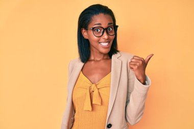 Genç Afro-Amerikan kadın, iş kıyafetleri içinde mutlu yüzle gülümseyen ve başparmağıyla yüzünü gösteren.. 