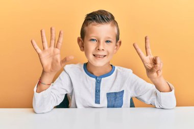 Sevimli beyaz çocuk, günlük giysiler içinde masada oturuyor ve yedinci parmaklarını göstererek gülümsüyor ve mutlu.. 