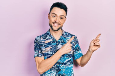 Gündelik kıyafetler giyen genç İspanyol adam gülümseyip kameraya bakıyor ve iki eliyle ve parmaklarıyla kenarı işaret ediyor.. 