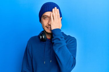 Eşofman altı ve kulaklık giyen İspanyol genç bir adam bir gözünü eliyle kapatıyor, yüzünde kendine güvenen bir gülümseme ve sürpriz bir duygu var.. 