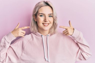 Yüzünde mutlu ve havalı bir gülümsemeyle parmaklarıyla dudaklarını gösteren genç sarışın kız. diş gösterme. 