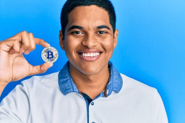 Sanal para birimini elinde tutan yakışıklı İspanyol adam pozitif ve mutlu görünüyor ve kendine güvenen bir gülümsemeyle dişlerini gösteriyor. 