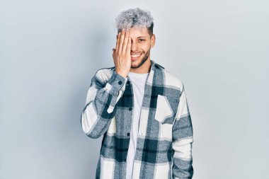 Modern boyalı saçları olan genç İspanyol bir adam gündelik gömlek giyiyor bir gözünü eliyle kapatıyor, yüzünde kendine güvenen bir gülümseme ve şaşırtıcı bir duygu var.. 