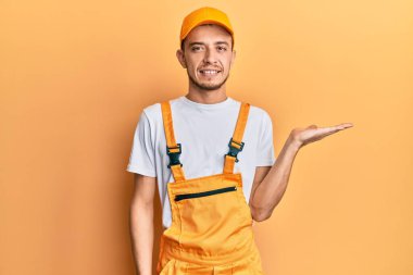 Tamirci üniforması giyen İspanyol genç adam neşeli bir şekilde gülümsüyor ve avuç içi kameraya bakıyor.. 