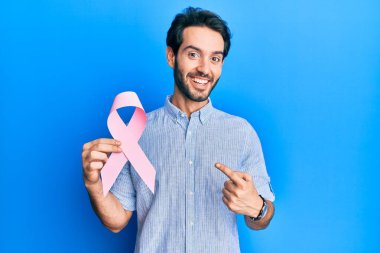 Genç İspanyol adam pembe kanser kurdelesi tutuyor gülümsüyor mutlu bir şekilde eli ve parmağı ile işaret ediyor 