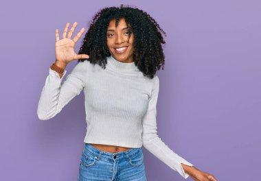 Genç Afro-Amerikan kızı günlük giysiler giyip beş numaralı parmaklarıyla işaret ederken kendinden emin ve mutlu bir şekilde gülümsüyor.. 