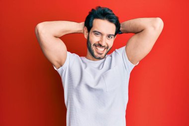 Latin asıllı genç adam, günlük beyaz tişört giyiyor, rahatlıyor ve esniyor, kolları ve elleri başının ve boynunun arkasında mutlu bir şekilde gülümsüyor. 