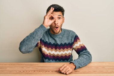 Masada oturan sıradan süveter giyen yakışıklı genç adam şaşırmış yüz ifadesiyle şaşırmış, parmaklarının arasından bakan bir jest yapıyor. İnançsız ifade. 