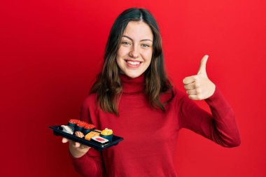 Genç esmer kadın bir tabak suşi gösteriyor mutlu ve pozitif gülümsüyor, başparmağıyla mükemmel ve onay işareti yapıyor. 