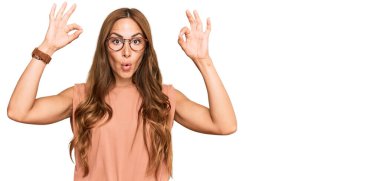 Gündelik kıyafetler ve gözlük takan genç İspanyol kadın şaşırmış ve şok olmuş bir şekilde parmaklarıyla onay sembolü çiziyor. çılgın ifade 