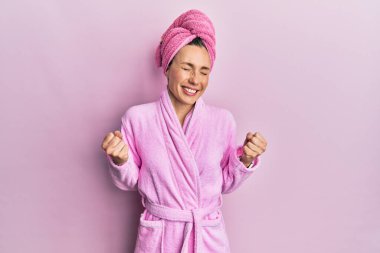 Banyo havlusu şapkası ve bornoz giyen genç sarışın kadın çok mutlu ve heyecanlı bir şekilde kollarını kaldırıp, gülümseyerek ve başarı çığlıkları atarak zafer jesti yapıyor. kutlama konsepti. 