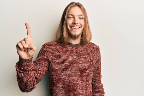 Красивый белый мужчина с длинными волосами, в обычном зимнем свитере, улыбается с идеей или вопросом, показывая пальцем вверх со счастливым лицом, номер один 
