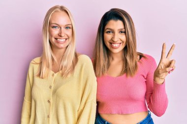 Pembe arka planda yan yana duran iki arkadaş iki numarayı gösterip işaret ederken kendinden emin ve mutlu bir şekilde gülümsüyor.. 