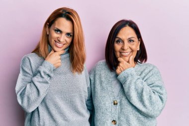 Latin anne ve kız günlük kıyafetler giyip kameraya bakarak kollarını kavuşturup gülümsüyor ve elleri çenesinde yükseliyor. Olumlu düşünmek. 